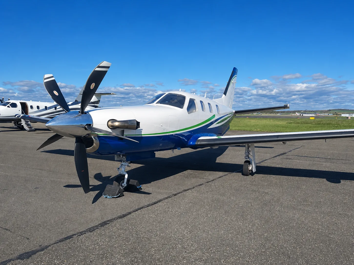 A white and blue single-engine turboprop aircraft with a green stripe is parked on an asphalt tarmac under a blue sky with clouds.