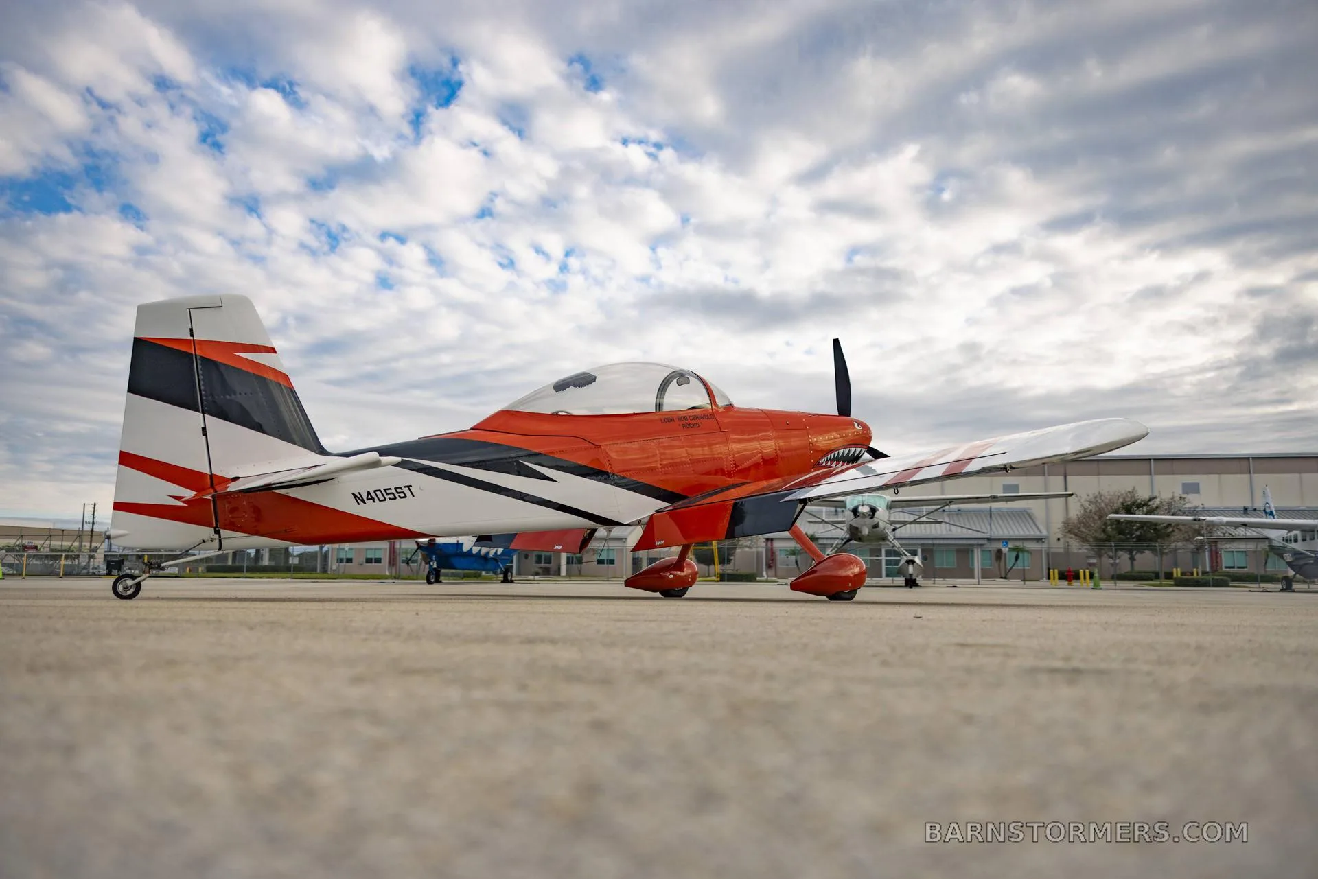 A side view of an orange, white, and black single-engine aircraft with a shark mouth paint scheme, parked on an airport tarmac under a cloudy sky.
