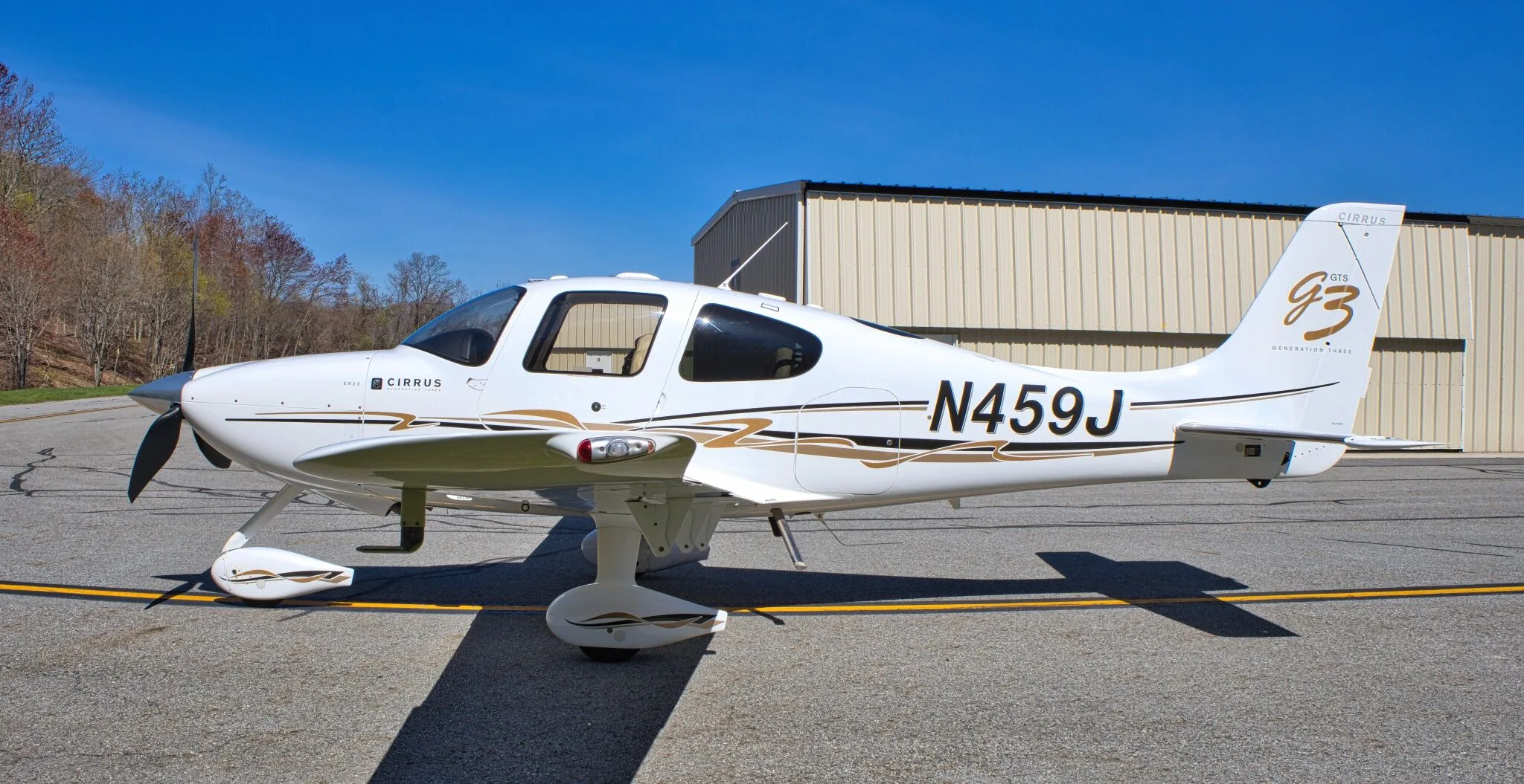 A white Cirrus SR22 GTS Generation Three aircraft (N459J) with gold and black stripes is parked on an airfield next to a hangar.