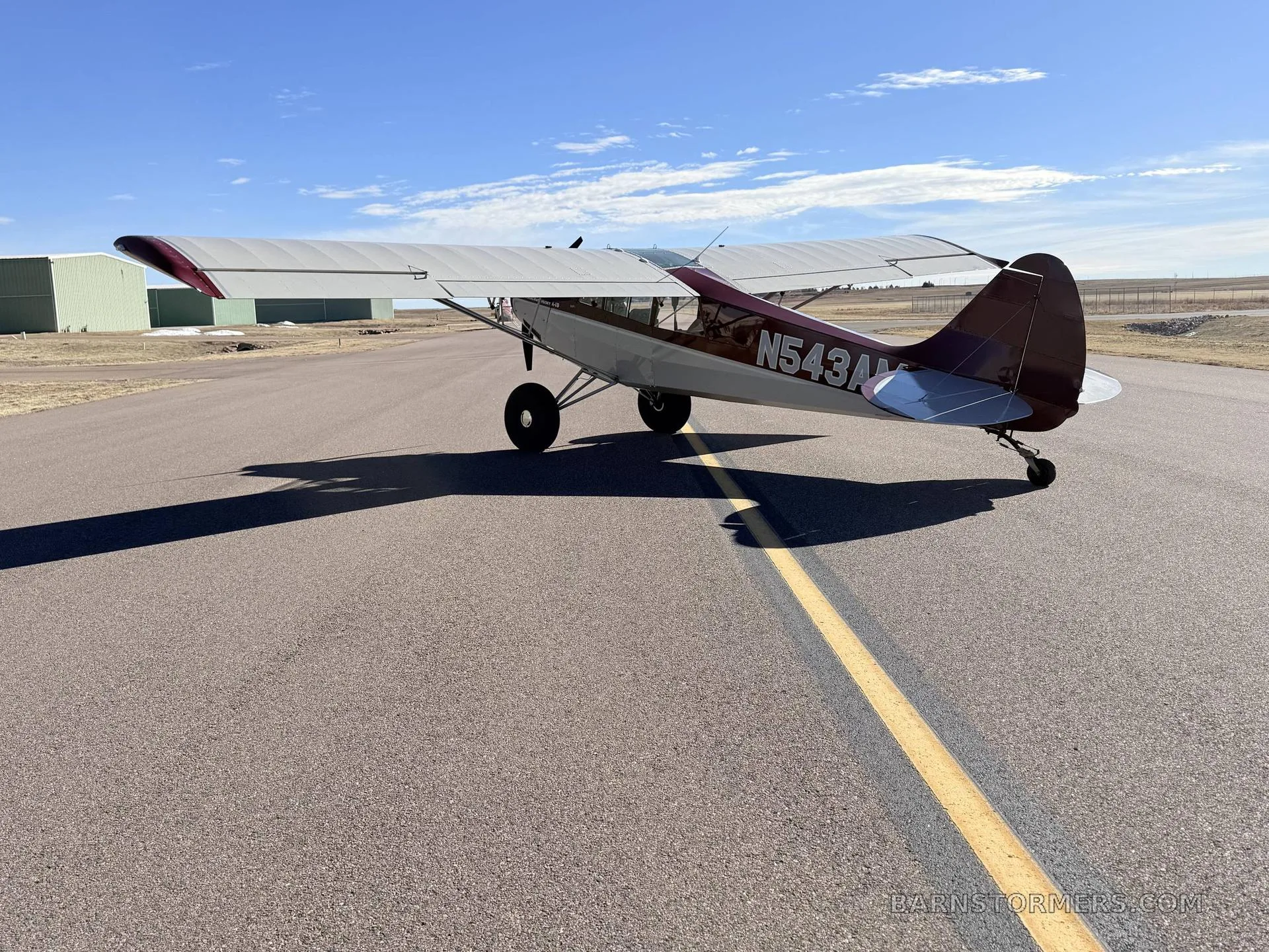 A red and white Aviat Husky A-1B aircraft with large tires is seen from the rear on an airfield, casting a long shadow.