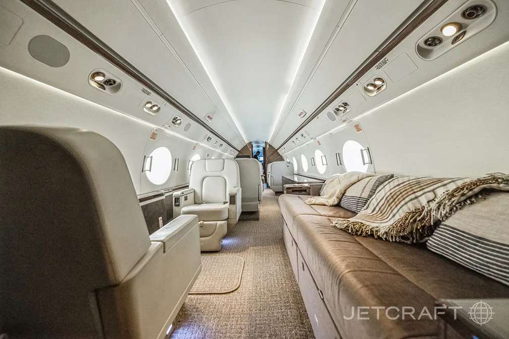 The spacious interior cabin of a private jet features light-colored seats, a long brown leather divan with blankets, and large circular windows.