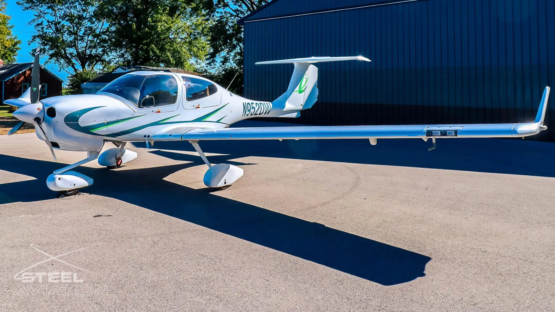 White Diamond DA40 aircraft with green accents and registration N952DW parked on an asphalt surface next to a dark blue hangar.