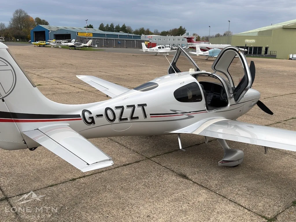 A silver Cirrus SR22T aircraft with registration G-OZZT and open doors, viewed from the rear-side on an airfield.