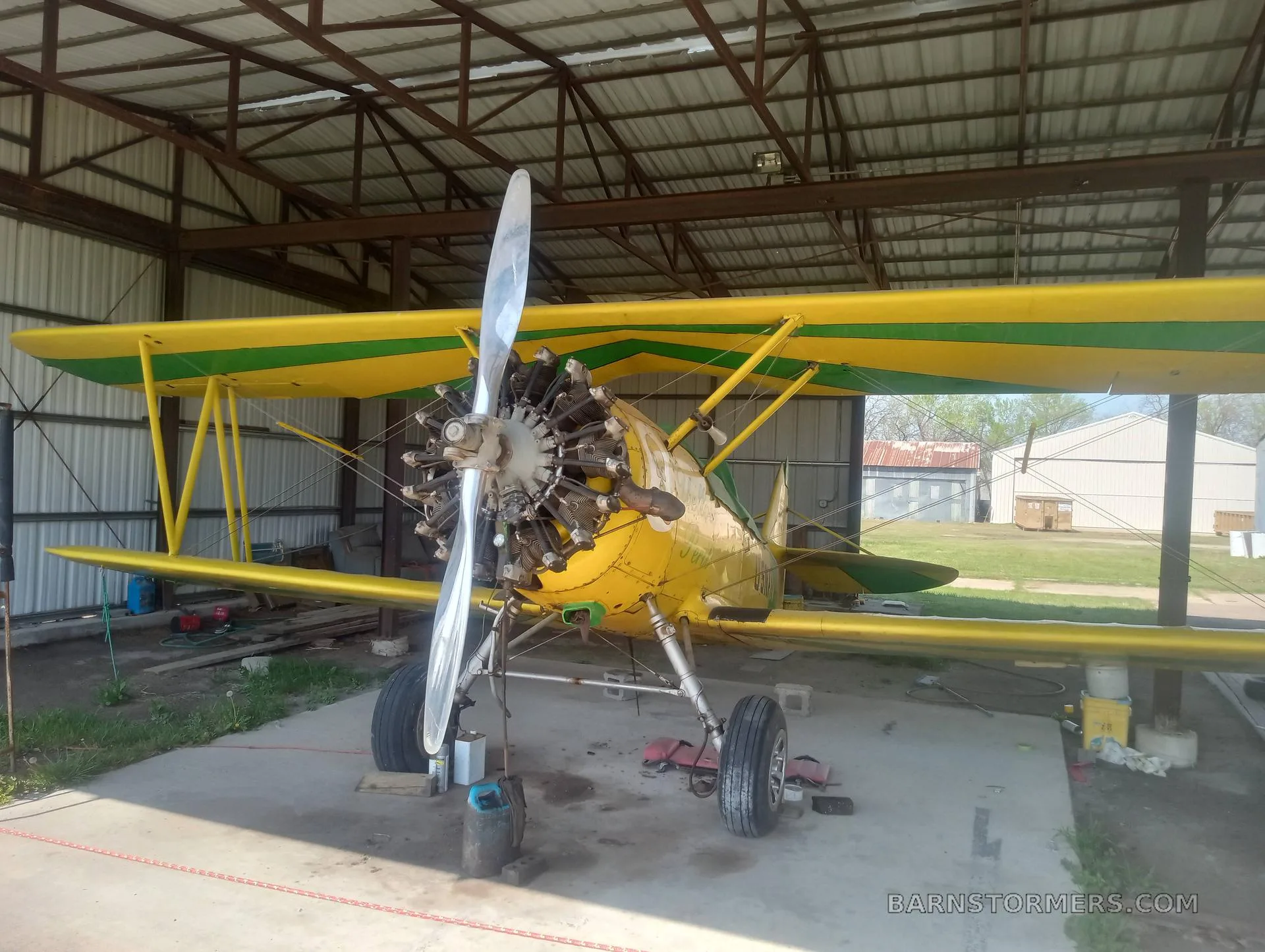 A yellow and green N3N biplane with an exposed radial engine is parked inside a hangar.