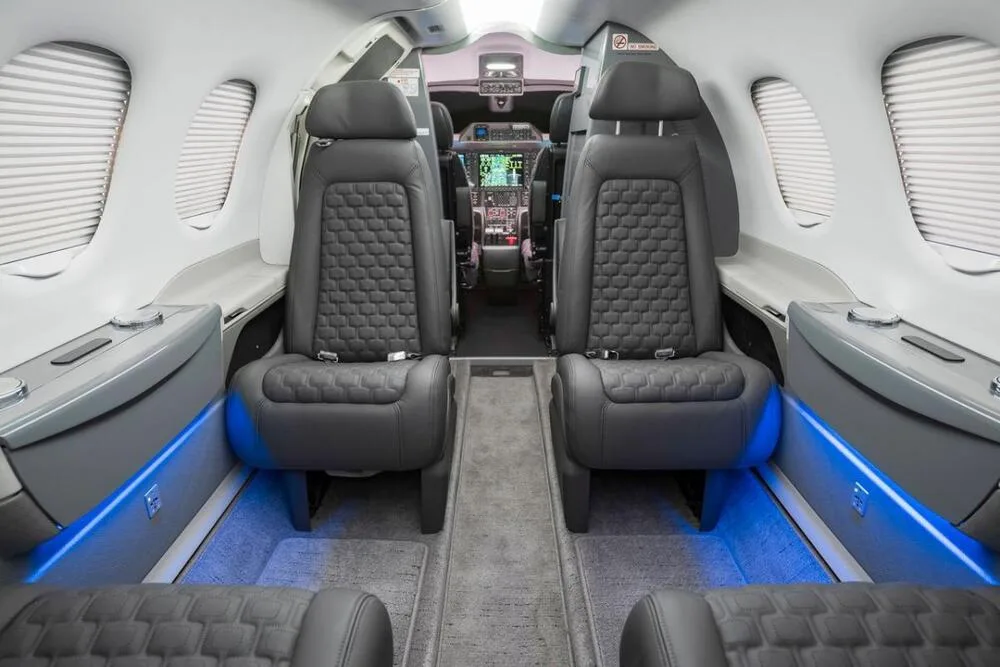 The interior cabin of an Embraer Phenom 100 with dark gray quilted seats and blue accent lighting, looking towards the cockpit.