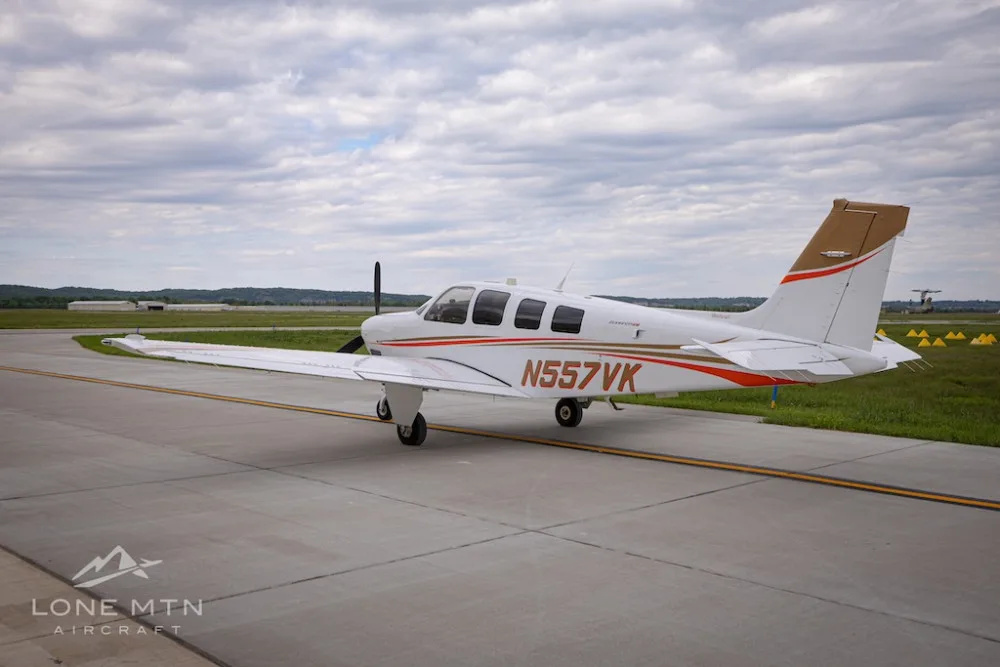 A rear-side view of a white Beechcraft Bonanza G36 with orange and brown stripes, registration N557VK, parked on an airfield.