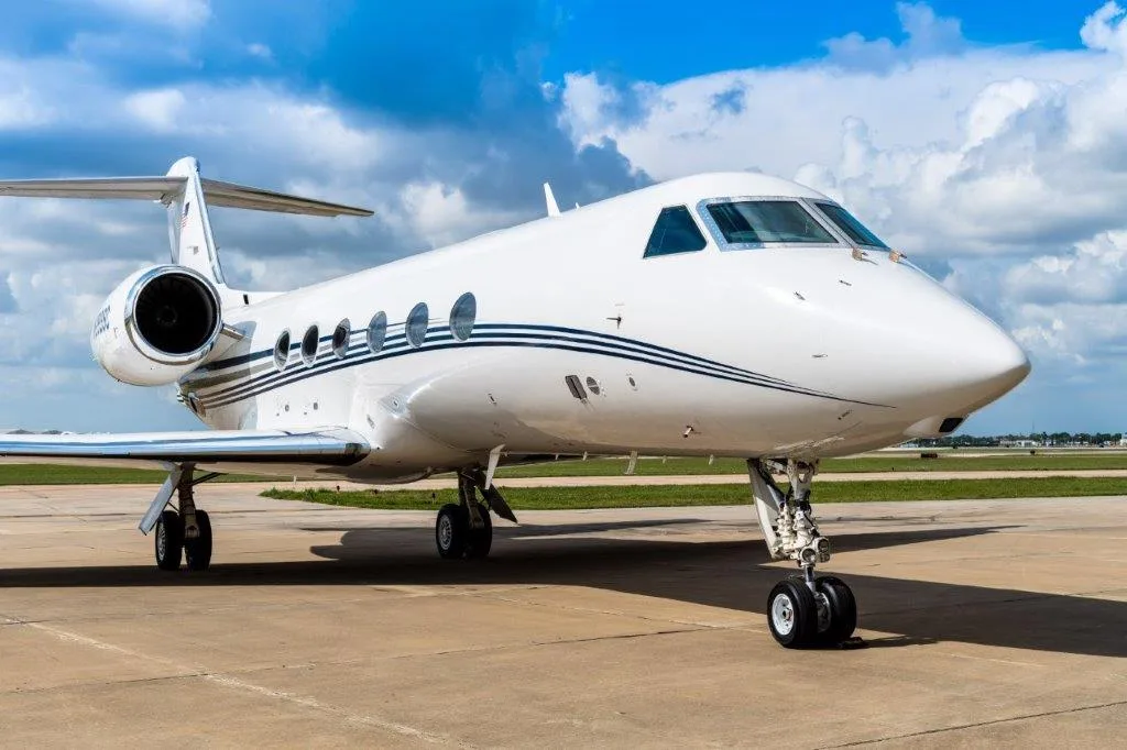 A white Gulfstream business jet with blue stripes is parked on an airport tarmac under a partly cloudy sky.