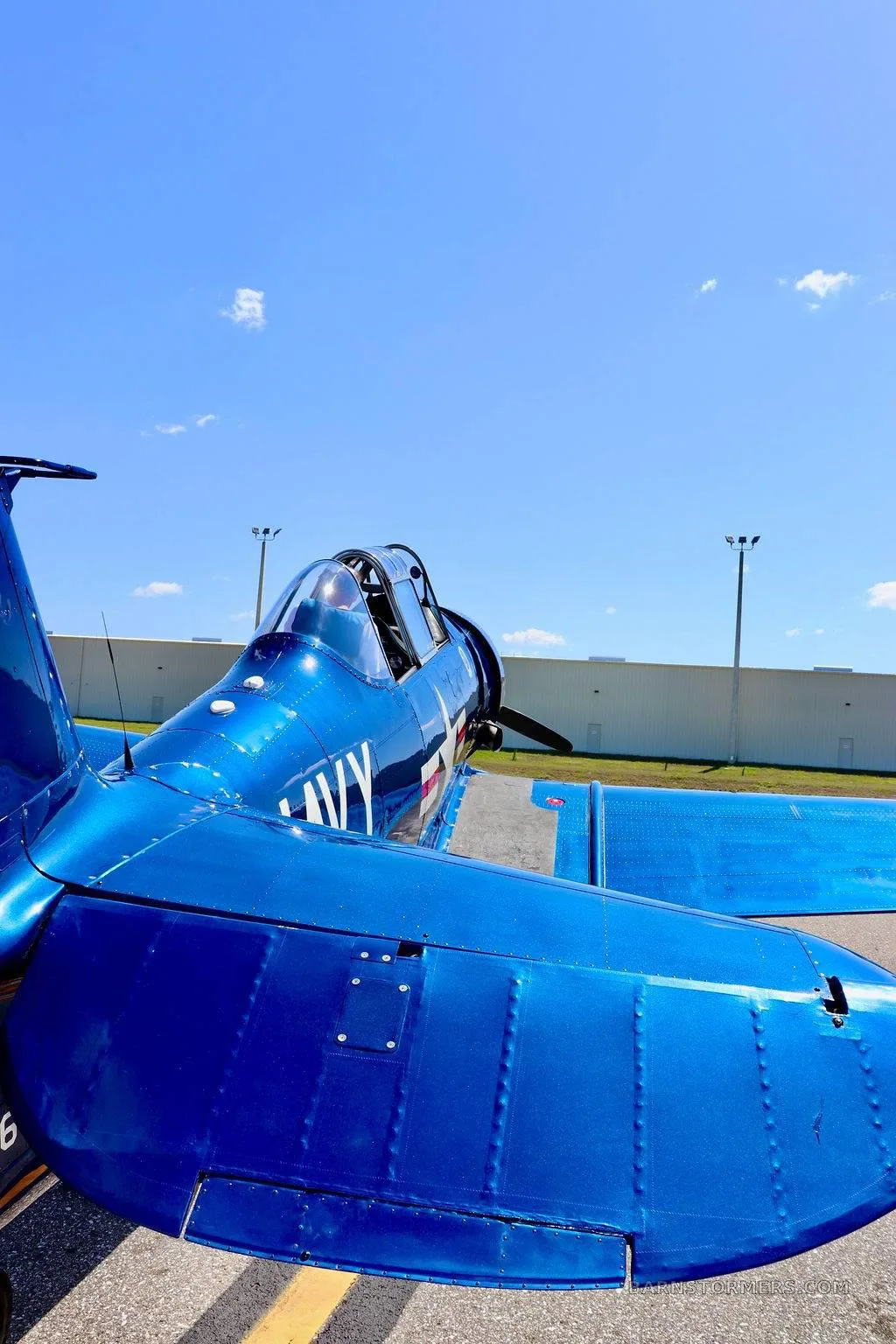 A close-up, rear-quarter view of a shiny dark blue Vought F4U Corsair aircraft with "NAVY" markings, parked on an airfield.