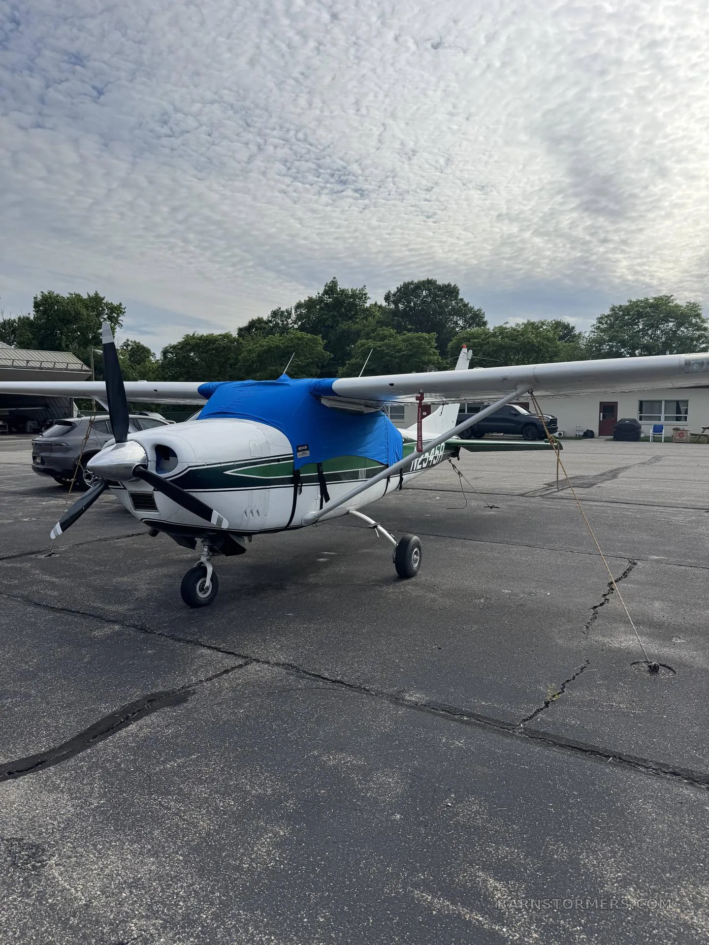 A white and green single-engine propeller aircraft with a blue engine cover is parked on an asphalt ramp, viewed from the front-right.