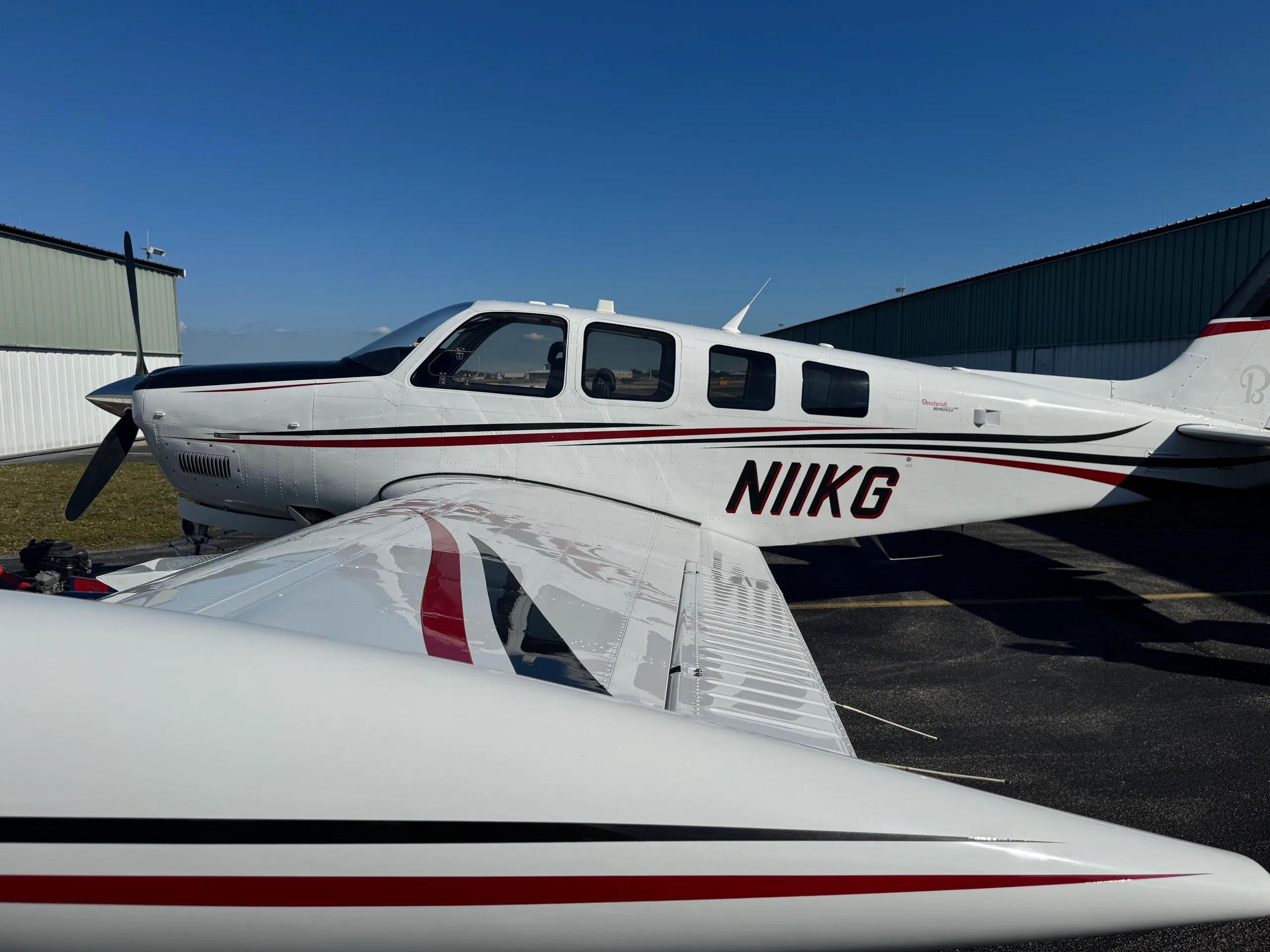 A close-up side view of the white Beechcraft Bonanza N11KG with black and red stripes on the tarmac.