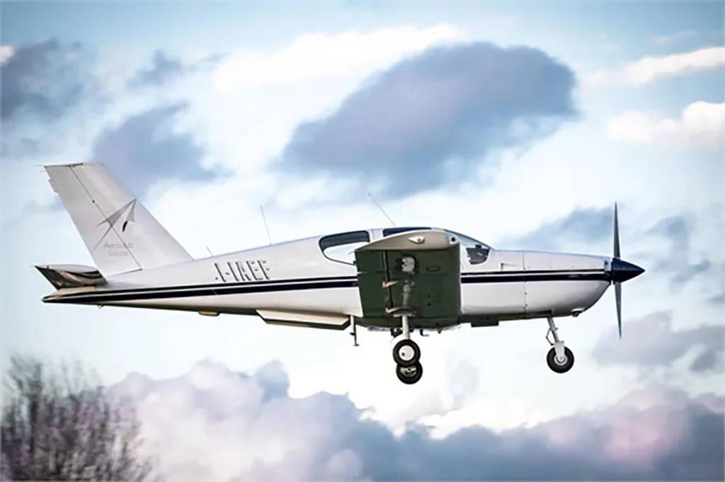 A white single-engine aircraft with black stripes and registration I-IAEF in flight with its landing gear extended against a cloudy sky.