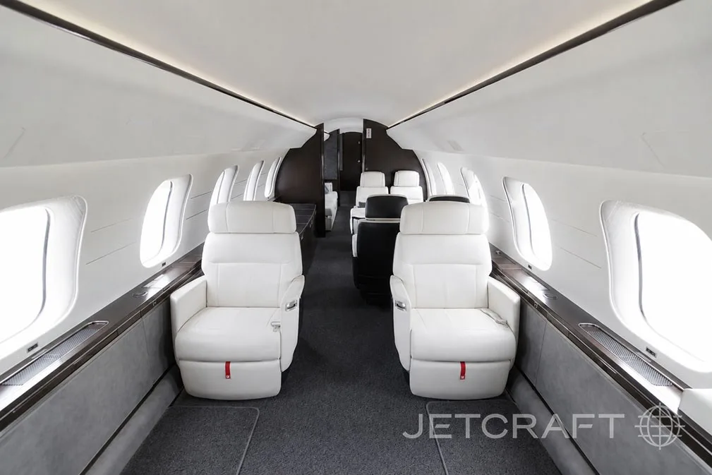 A view down the aisle of a luxurious aircraft cabin with white leather seats and large windows.