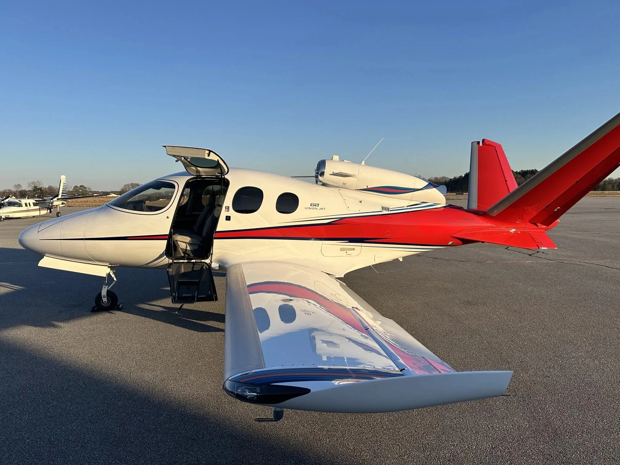 A white Cirrus Vision Jet with red and black accents, with its main door open, parked on an airfield.
