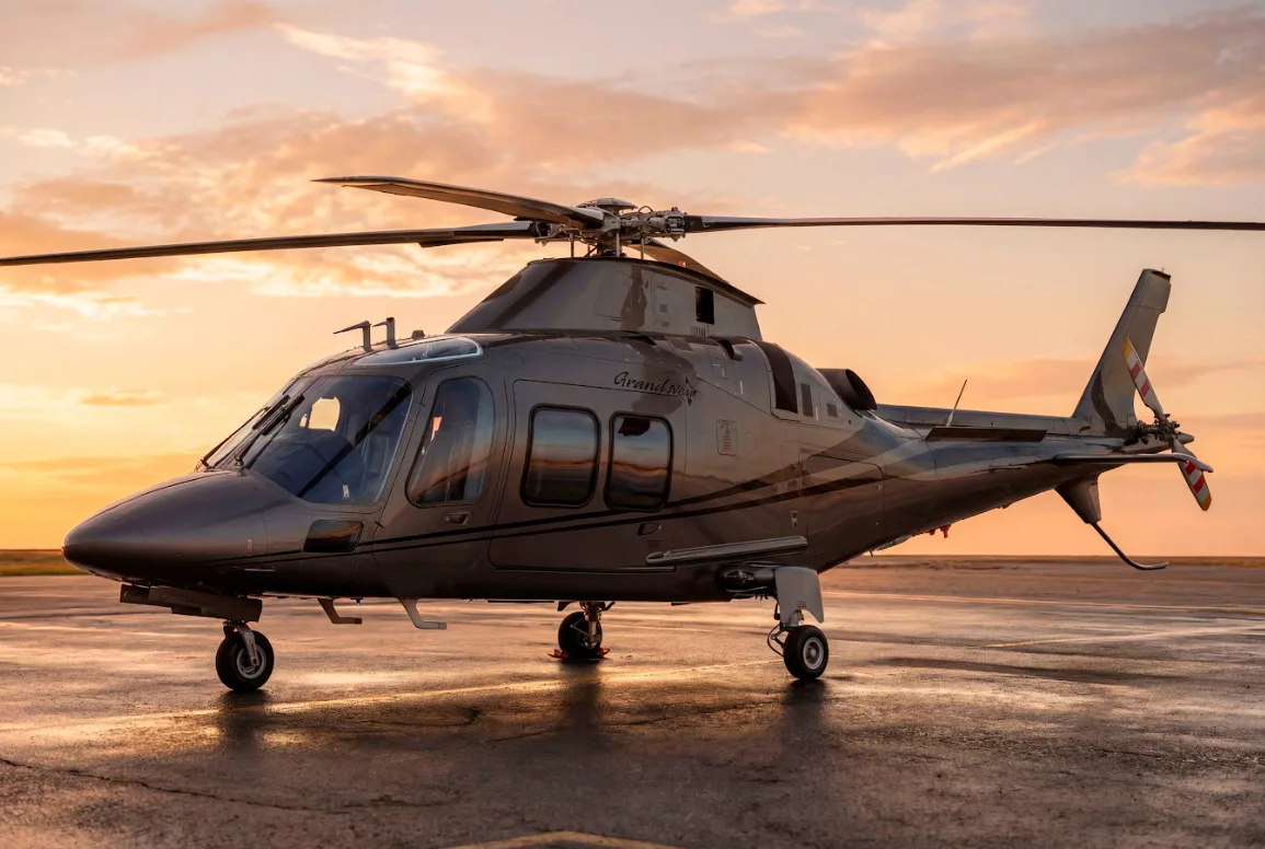 A grey AgustaWestland AW109 GrandNew helicopter is parked on a wet tarmac at sunset.