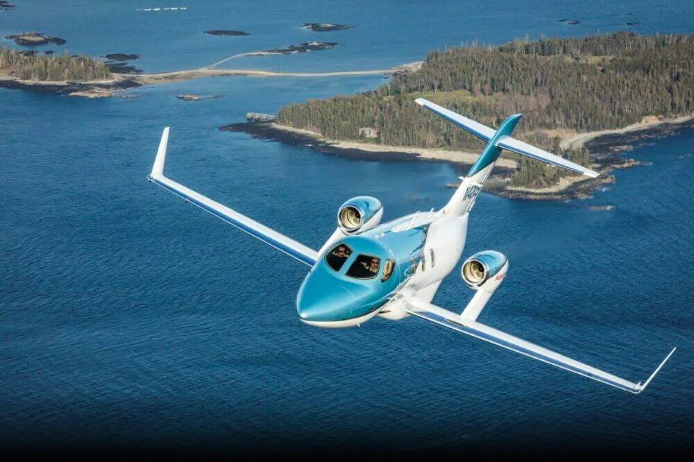 A light blue and white HondaJet (N420HJ) flies over a blue ocean with green, rocky islands.