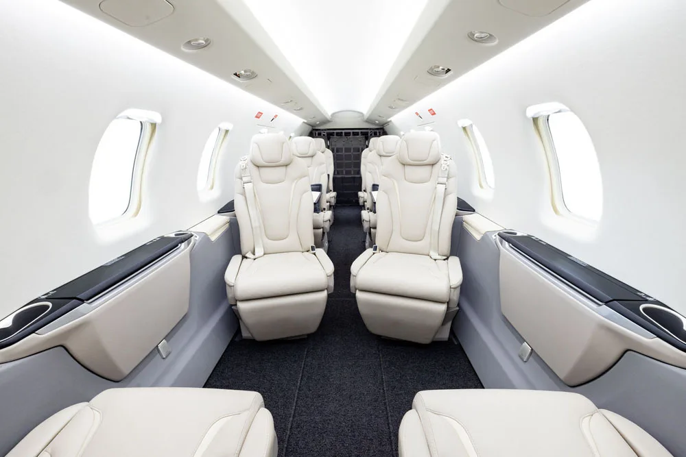 A view down the aisle of a private jet cabin with light beige leather seats and white walls.