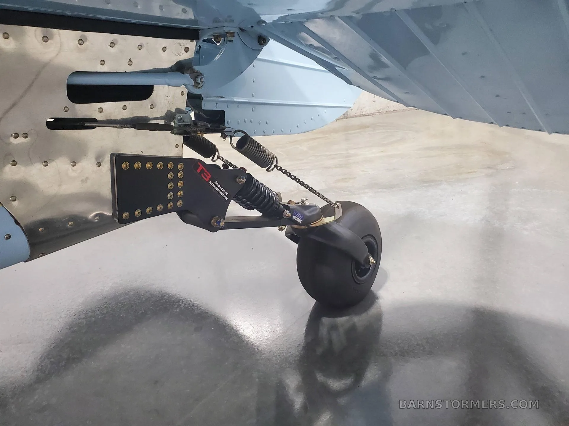 A close-up view of the tailwheel assembly of a light blue aircraft, showing the suspension and steering mechanism.
