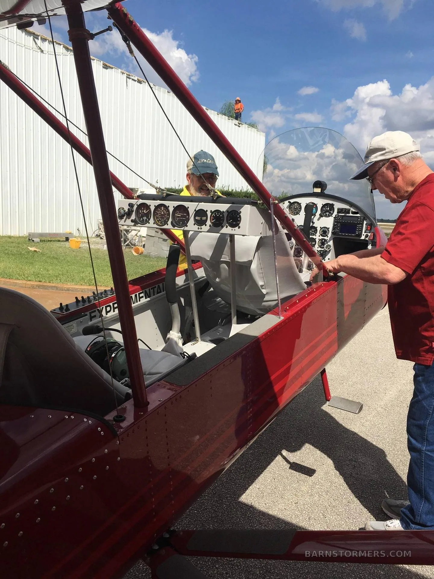 Two men inspect the open cockpit of a red experimental aircraft on an airfield.