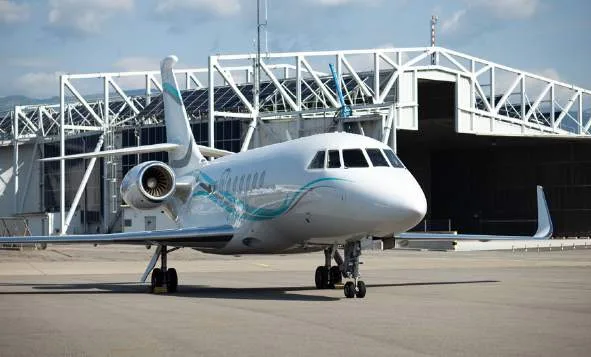 A white business jet with blue and teal stripes, viewed from the front, parked on an airfield in front of a large hangar.