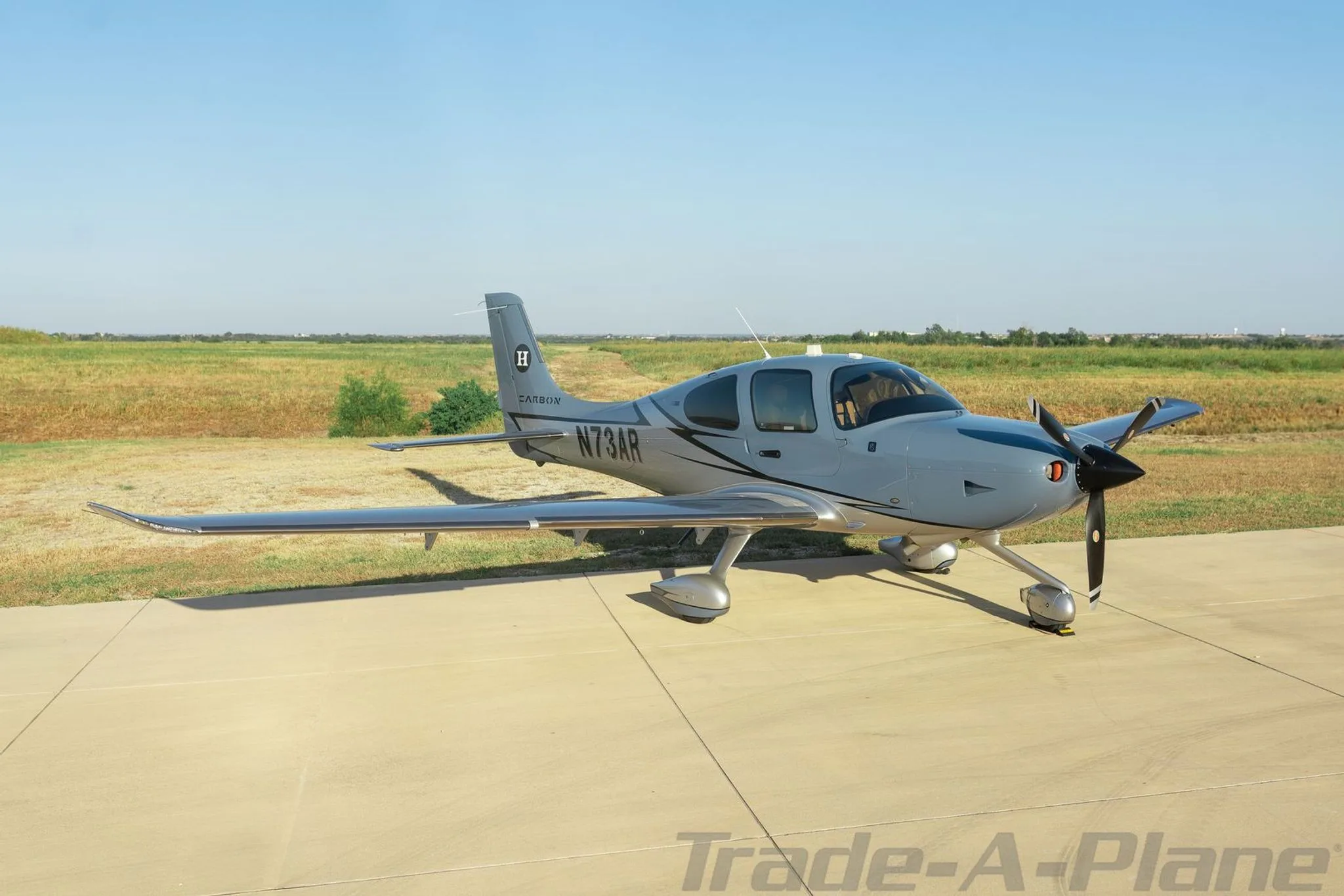 A side view of a grey Cirrus SR22 aircraft with registration N73AR parked on a tarmac next to a grassy field.