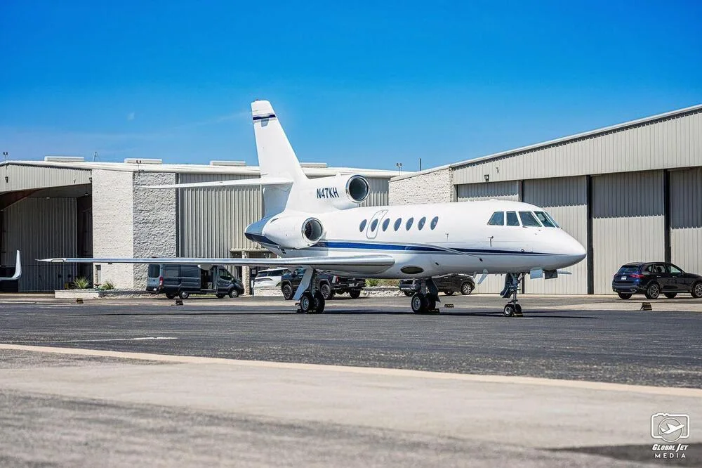 A white Dassault Falcon 900EX business jet with blue stripes, parked on an airfield with hangars in the background.
