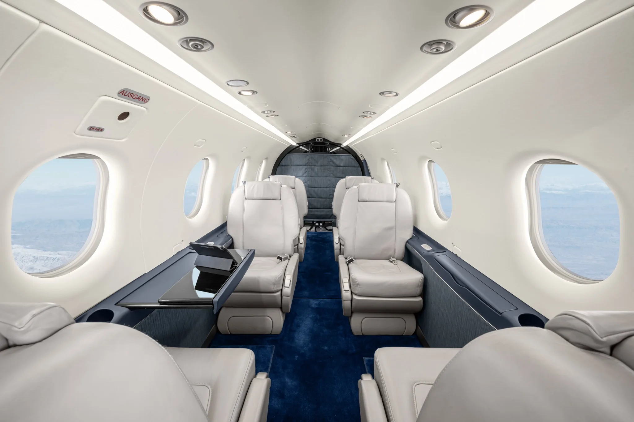 The aircraft's cabin interior features light grey leather seats, a blue carpet, and a small table with a tablet.