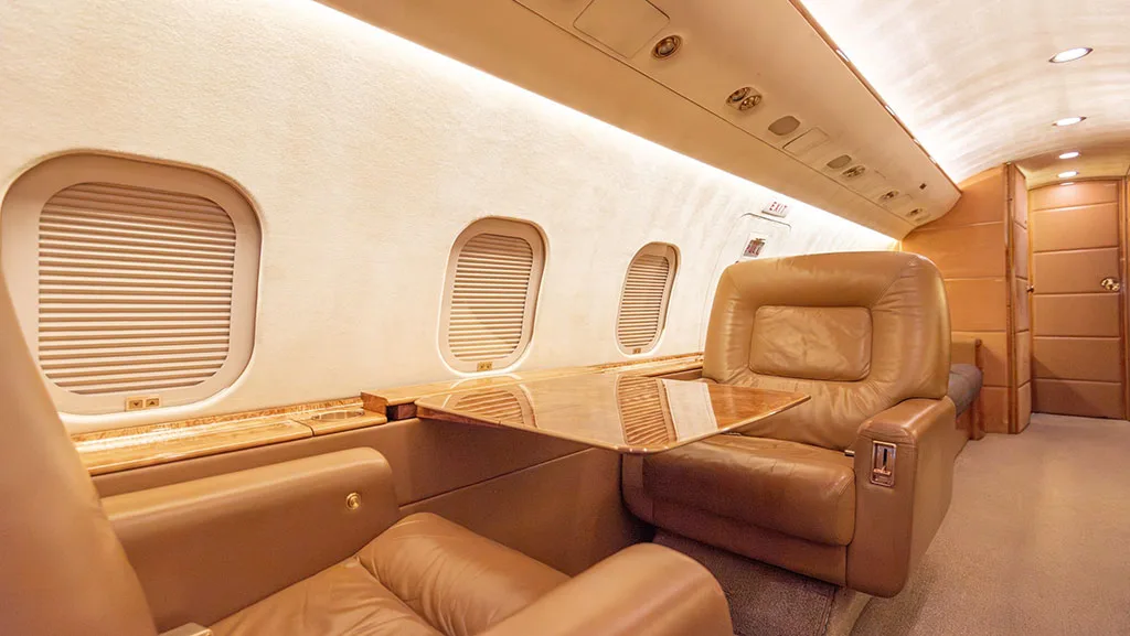 A close-up view of the private jet cabin with tan leather seats and a polished wooden table extended.