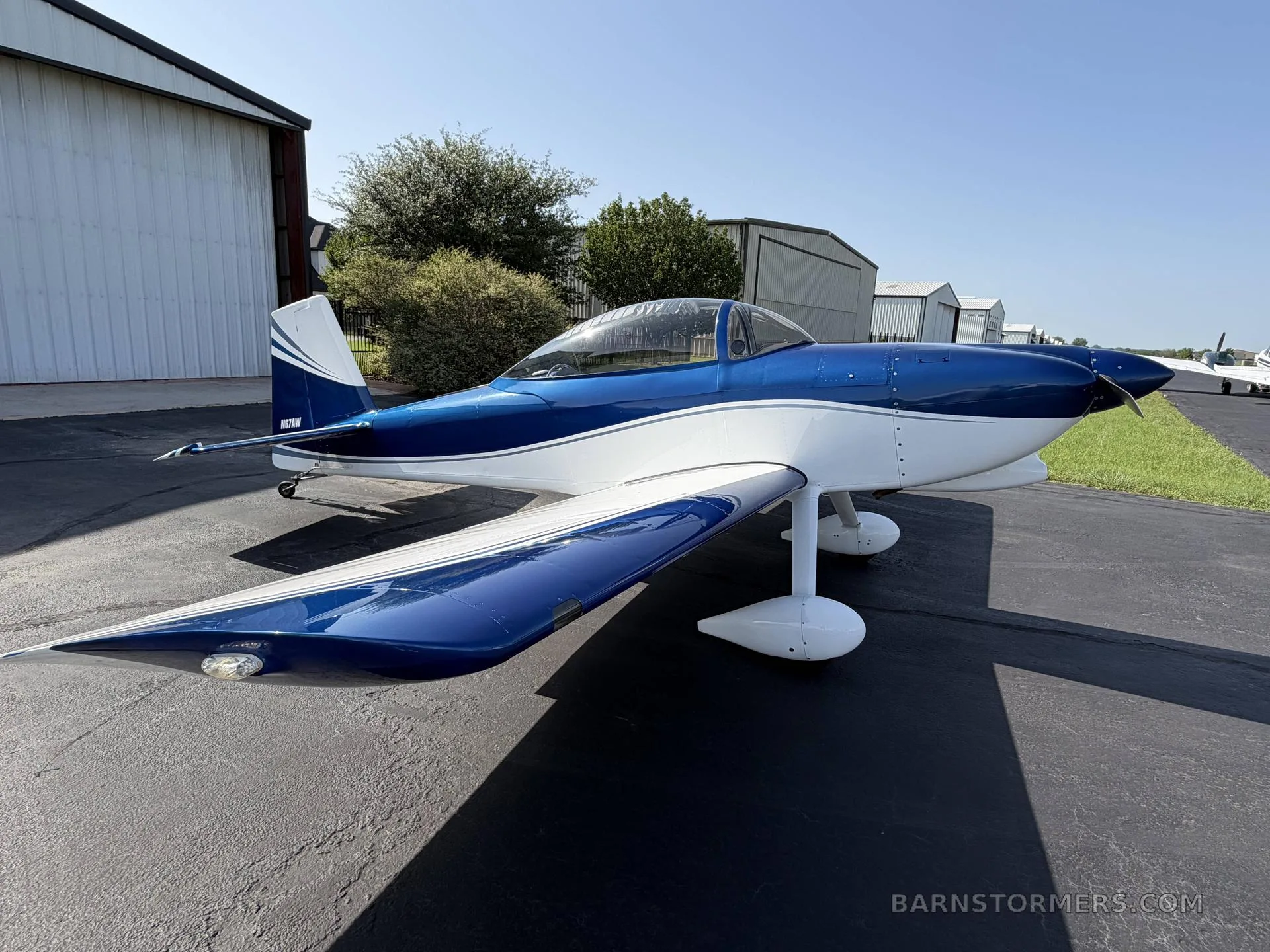 A blue and white single-engine aircraft is parked on a tarmac with hangars in the background on a sunny day.