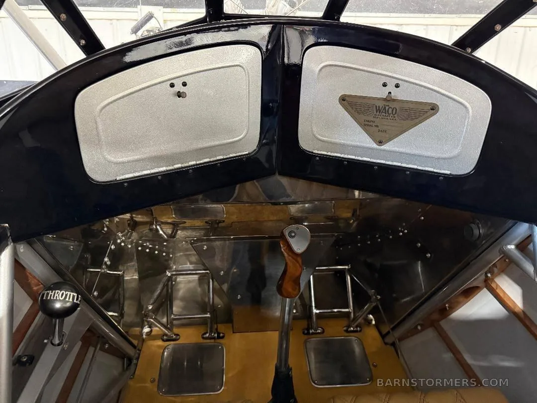 The front cockpit of a biplane shows a throttle, control stick, and a "WACO" manufacturer's plate.