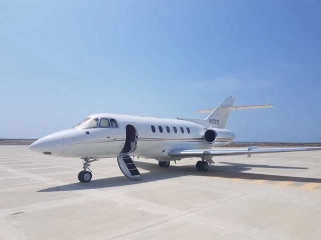 A white Hawker business jet with its main cabin door open and stairs extended, parked on a tarmac under a clear blue sky.