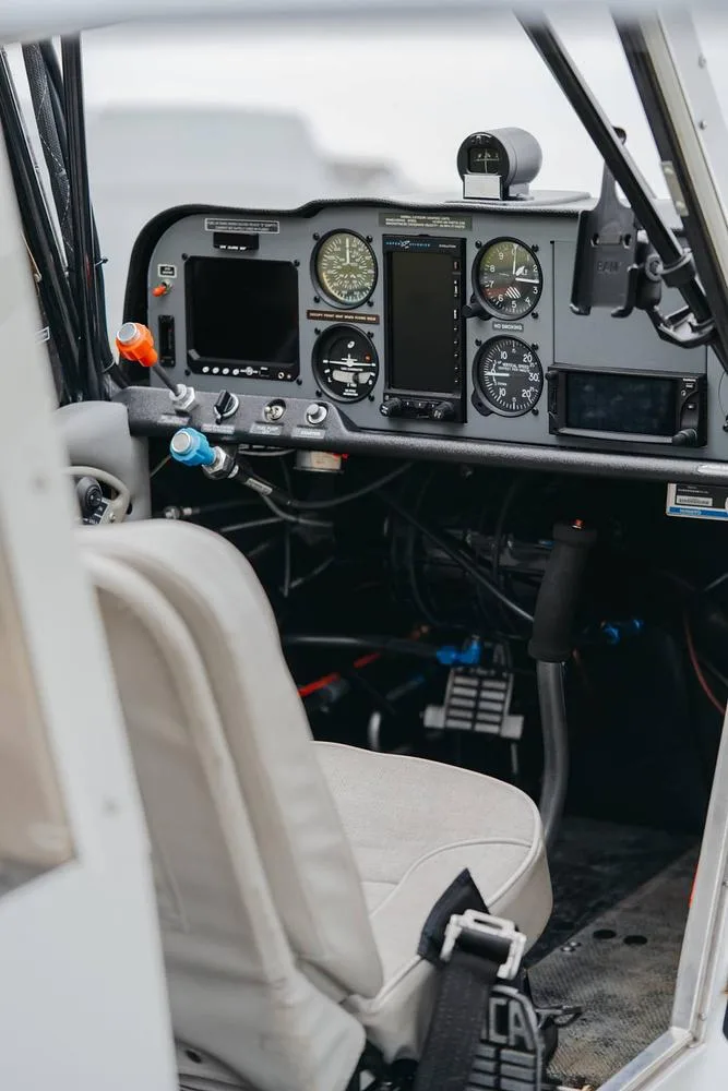 The cockpit of an aircraft, showing the instrument panel with gauges and screens, and a light-colored pilot's seat.