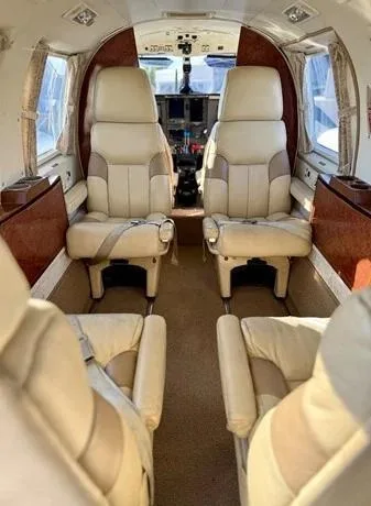 The aircraft's interior features light-colored leather seats and wood paneling, looking towards the cockpit.