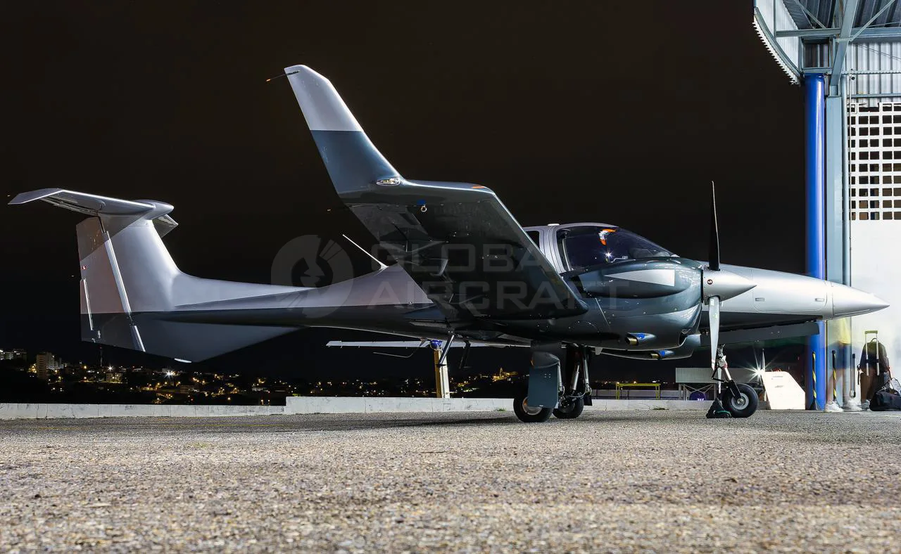 A silver and dark grey Diamond DA62 aircraft parked on a tarmac at night, with city lights in the background.