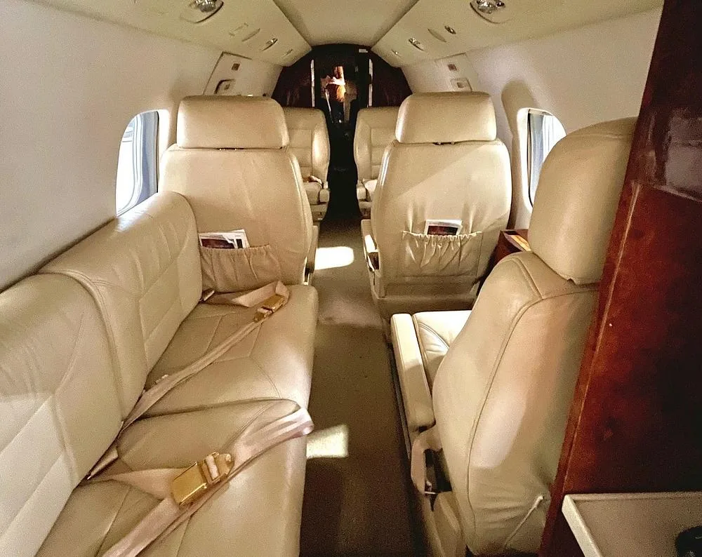 A long view of a private jet cabin with beige leather seating, including a bench seat on the left and individual seats on the right.