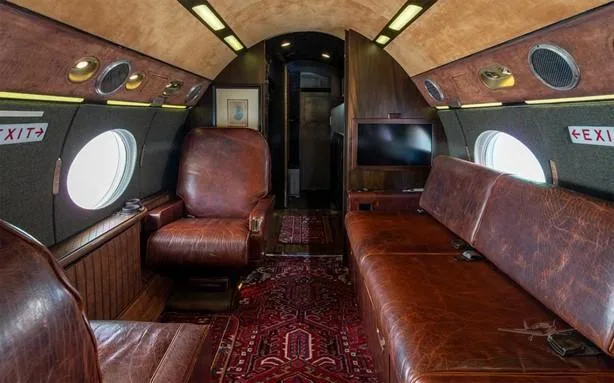 The aircraft cabin features a long brown leather sofa, a single brown leather armchair, and a patterned red rug.