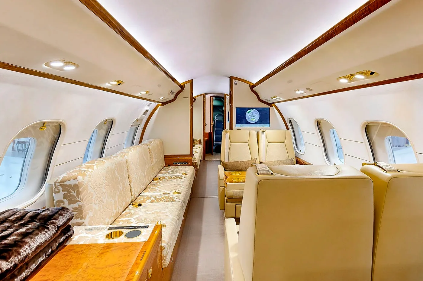 A spacious view of the private jet's cabin, showing a long patterned sofa, individual beige seats, and rich wood paneling.