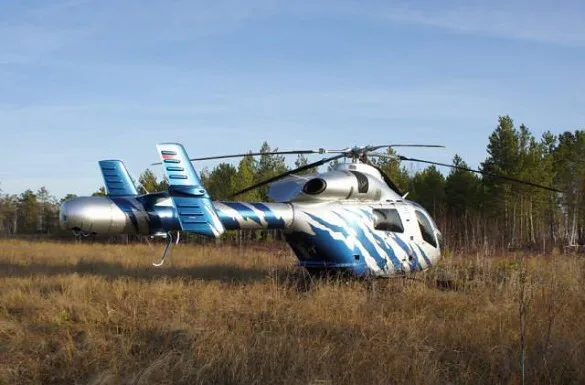 A silver and blue striped helicopter parked in a grassy field with a forest in the background.