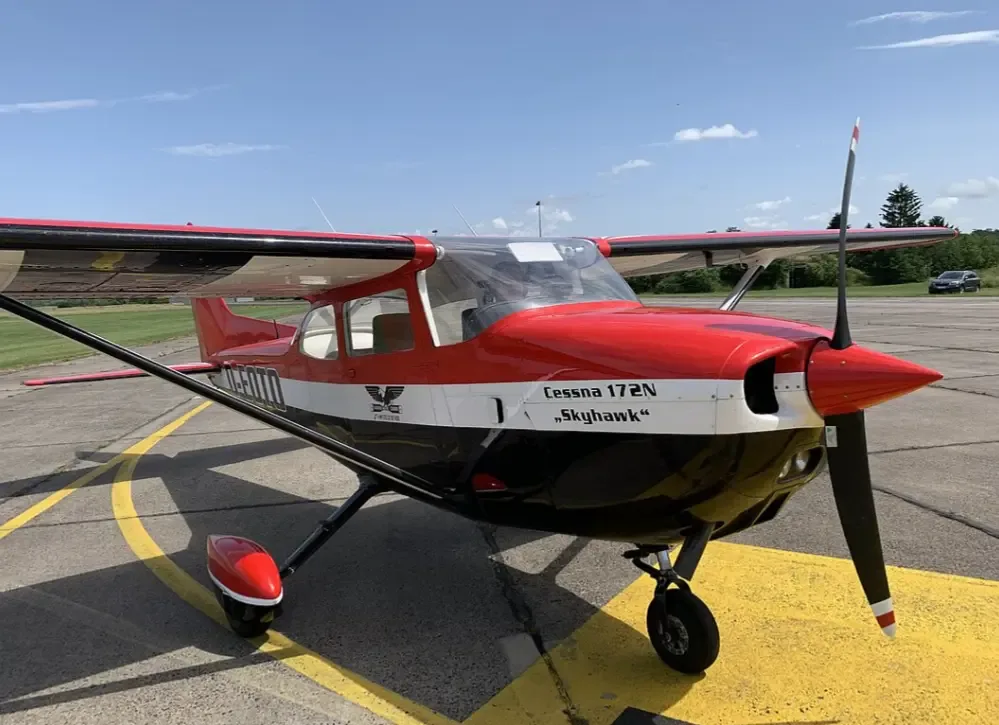 A red, white, and black Cessna 172N Skyhawk aircraft with a propeller, parked on an airport tarmac under a blue sky.