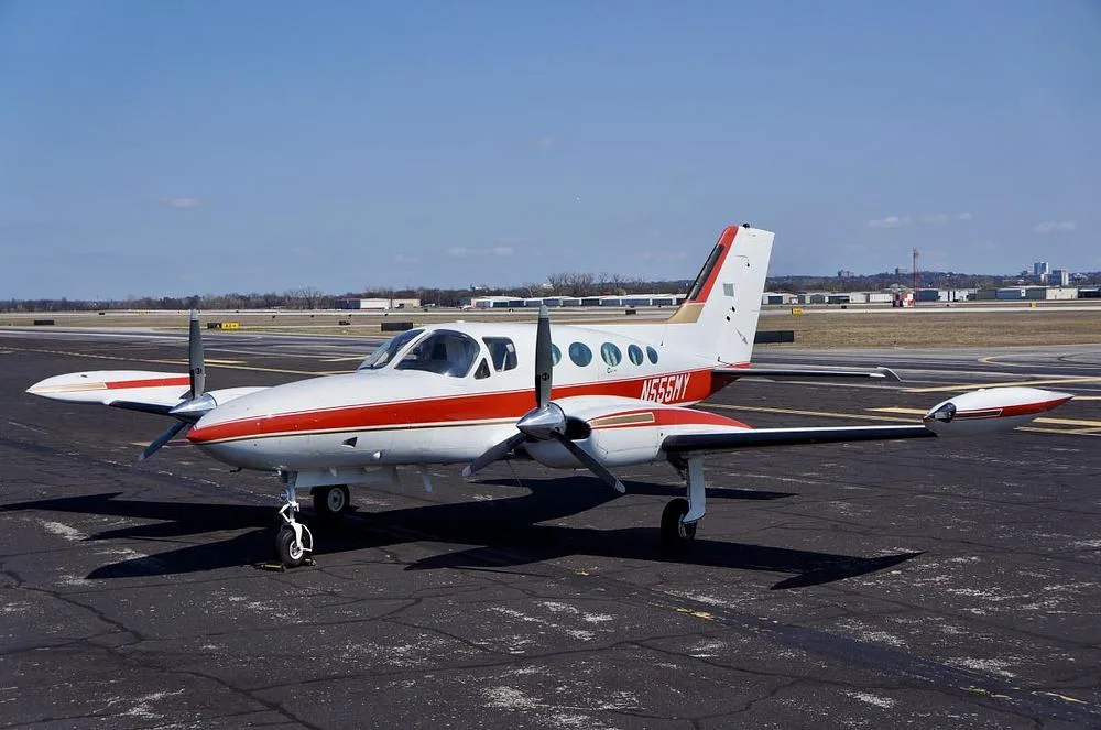 A white and red Cessna 400 series twin-engine aircraft, registration N555MY, is parked on an asphalt tarmac under a clear blue sky, viewed from the front-left.