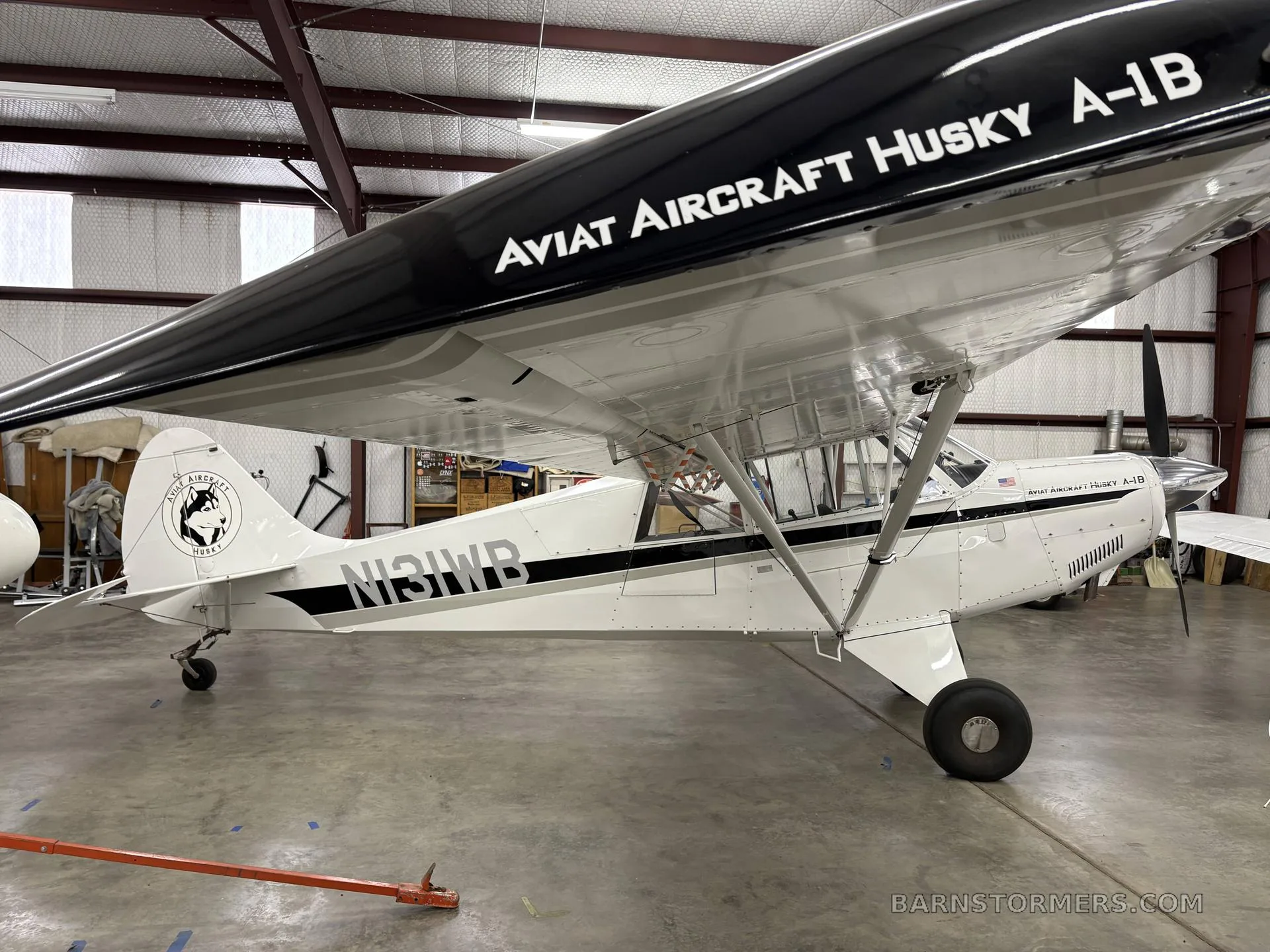 A white Aviat Aircraft Husky A-1B with black trim and tail number N131WB, parked in a hangar, viewed from the side.