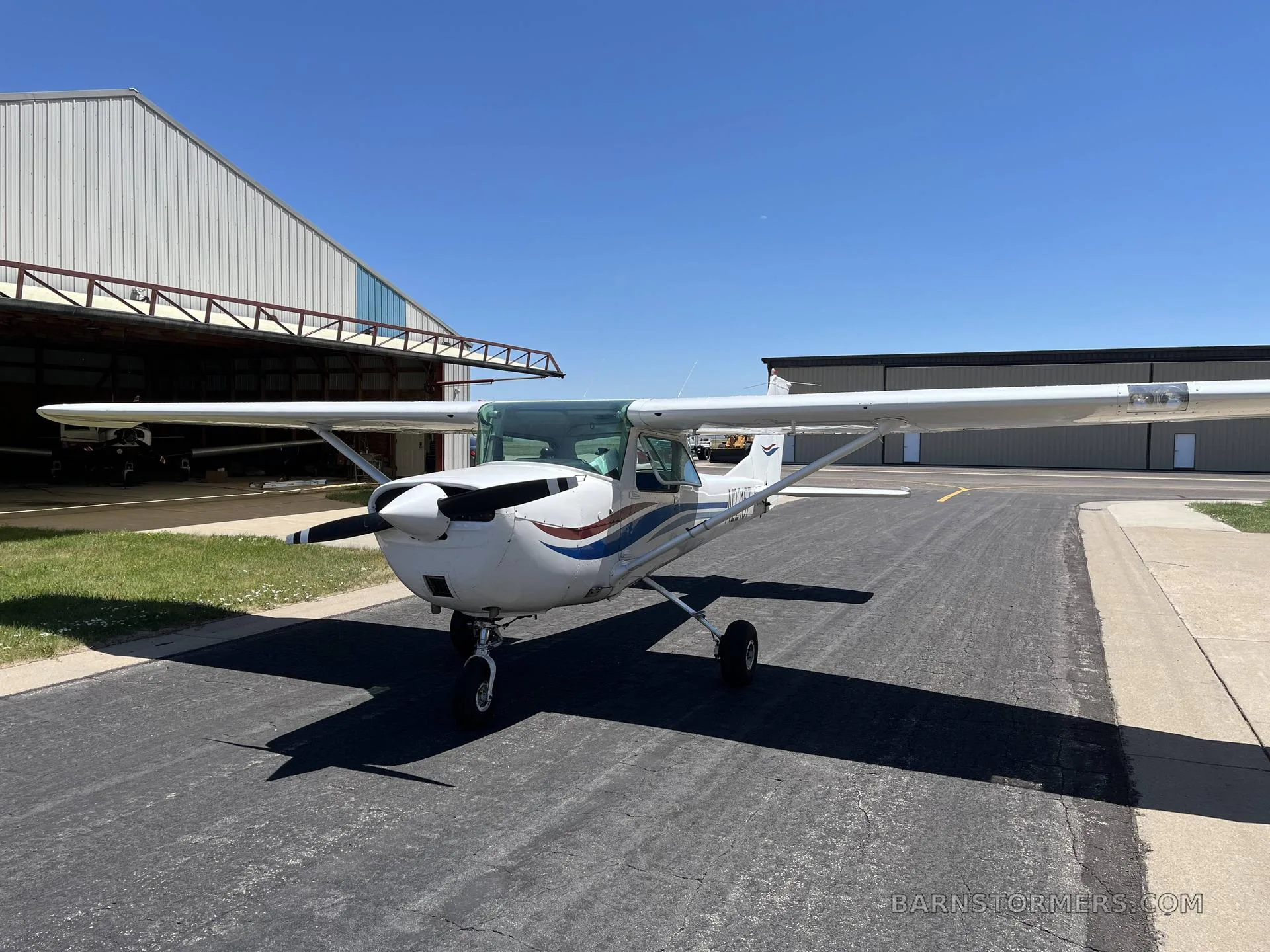A white Cessna 150/152 type aircraft with blue and red stripes parked on an asphalt ramp in front of a hangar on a sunny day.