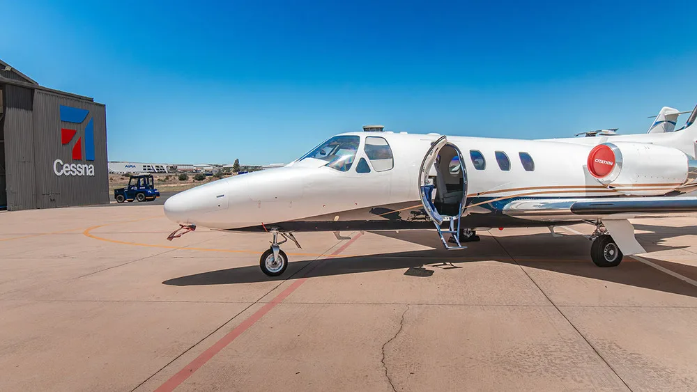 A white Cessna Citation jet with gold and blue stripes parked on an airfield, with its door open, next to a Cessna hangar.