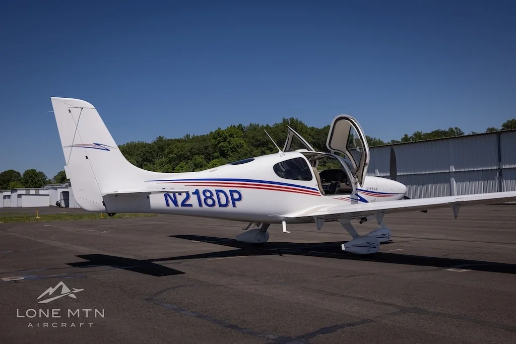 Side view of a white Cirrus aircraft (N218DP) with blue and red stripes, doors open, parked on an airfield.