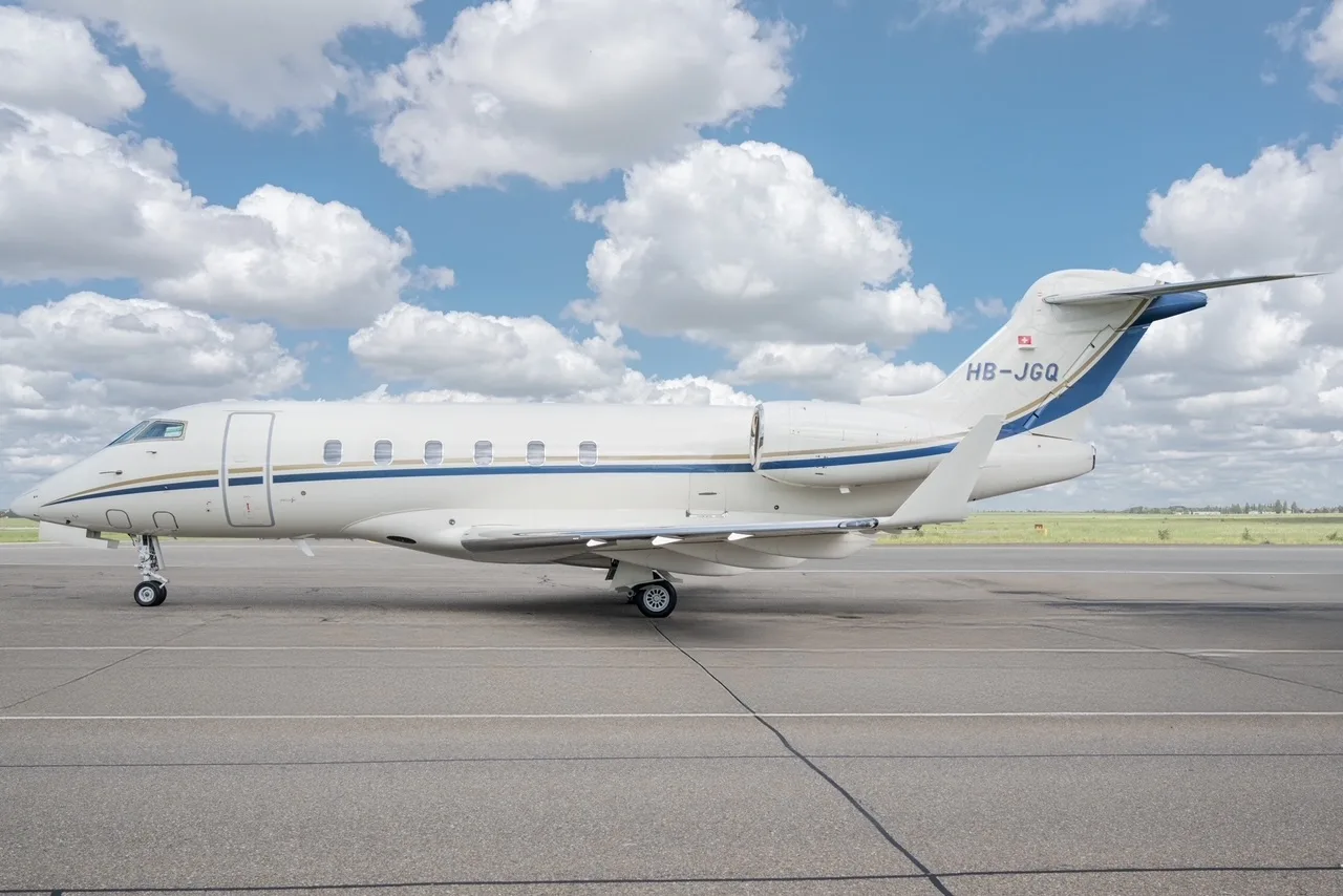 A white Bombardier Challenger 300 business jet with blue and gold stripes and tail number HB-JGQ parked on the tarmac.