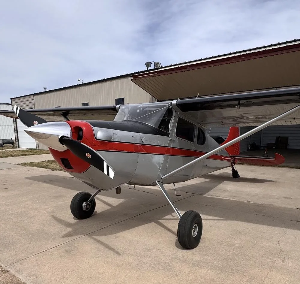A silver and red Cessna 170B aircraft parked on an airfield tarmac in front of a hangar.
