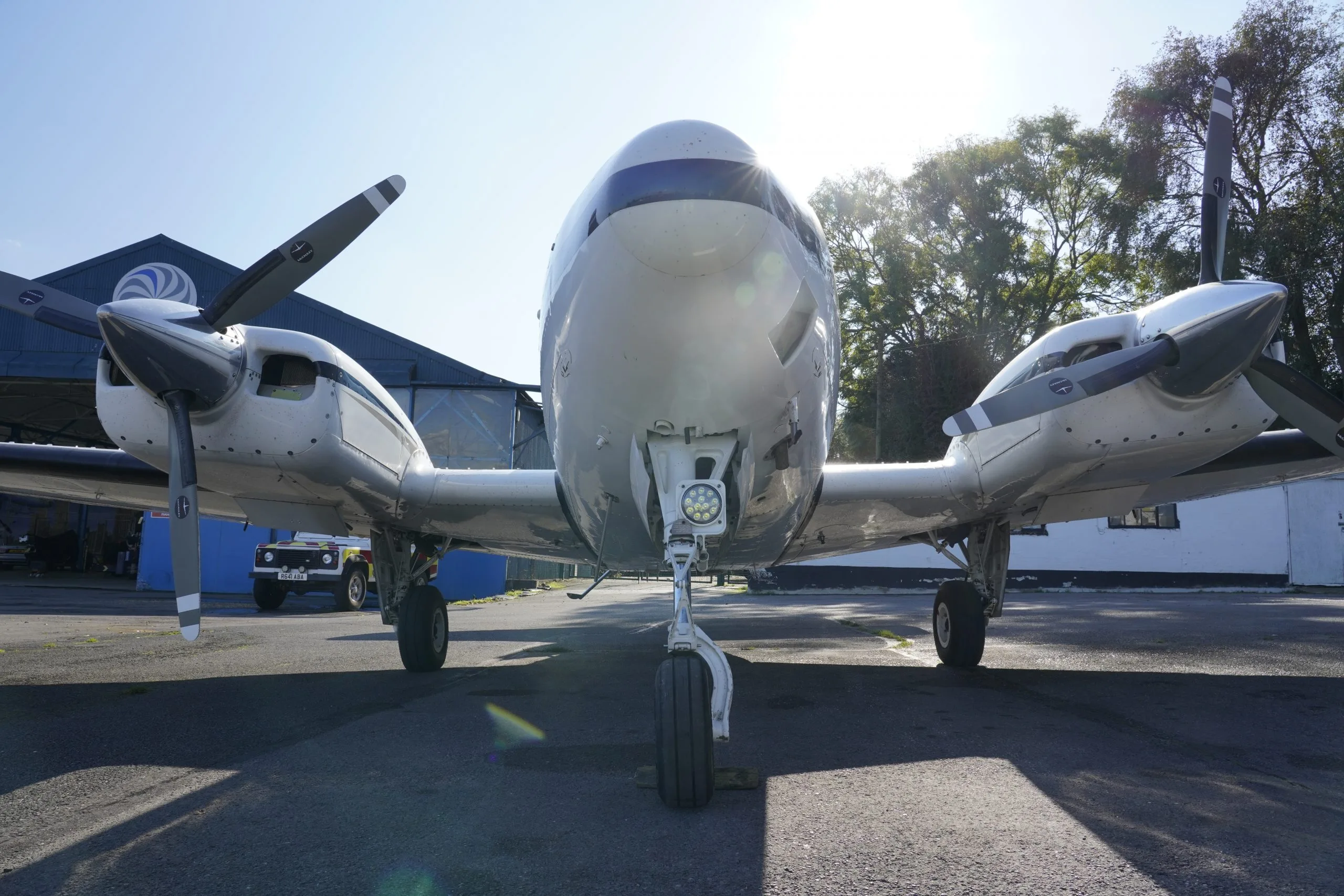 A head-on view of a white twin-engine propeller aircraft with blue stripes, parked on an asphalt apron.