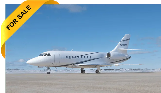 A white Dassault Falcon 900 business jet with dark blue stripes is parked on an airfield with snow in the background under a clear blue sky.