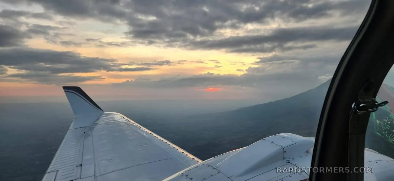 A view from the cockpit shows the white wing of an aircraft with black and red stripes, flying over a landscape with mountains at sunset.