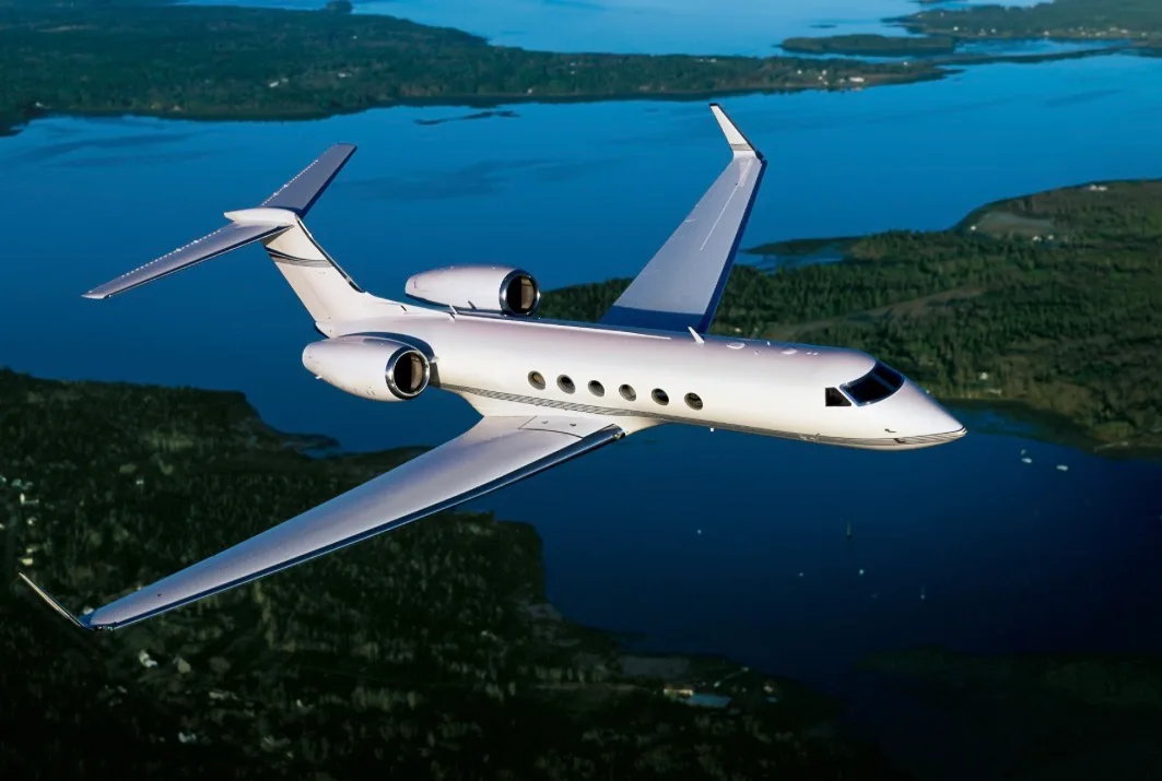 A white Gulfstream business jet with silver accents flies over a blue lake and green land.