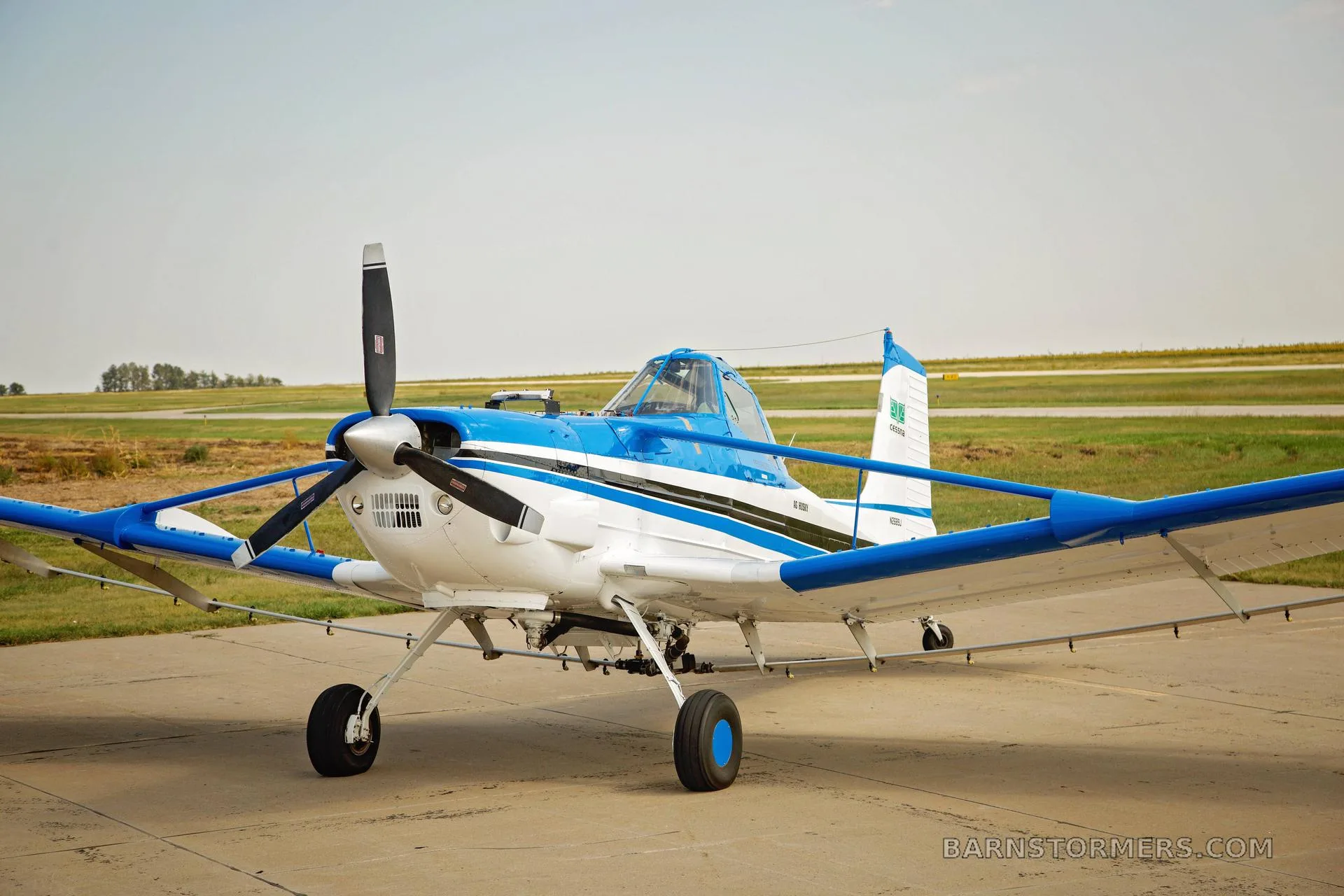 A white and blue agricultural aircraft, possibly a Cessna Ag Husky, is parked on a tarmac with a grassy field in the background.